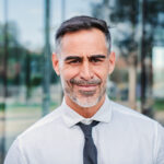 Close up portrait of successful mature caucasian business man smiling looking at camera standing outdoors at workplace with cheerful attitude. Front view of handsome executive employee wearing a suit. High quality photo